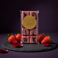Full packaged organic strawberry shell chocolate bar displayed with strawberries and rose petals on dark slate.